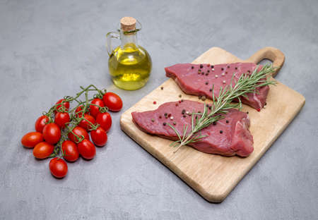 Steaks on a wooden board, next to a sprig of rosemary cherry tomatoes on a branch and a bottle of...