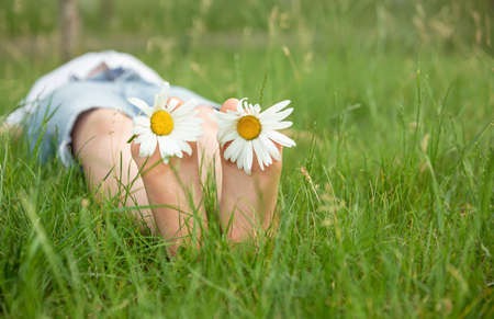Child with daisy lying in sunny meadow and relaxing in summer sunshine. Boy lying on green grass outdoors in park. Happy summer feet. Kid having fun at spring nature. Child feet with daisy flowerの写真素材