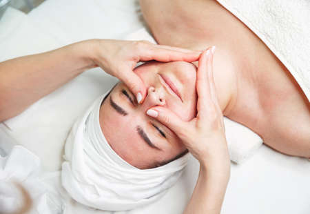Beautiful girl has a facial massage in a beauty clinic. Cosmetologist doing massage for woman. Facial massage beauty treatment. Young woman lying with closed eyes and having face and head massageの写真素材