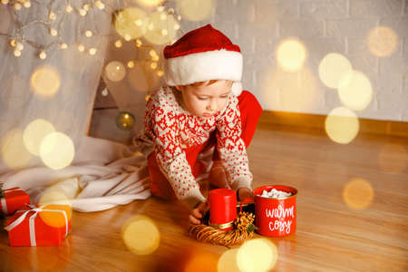 Adorable toddler in santa hat with gifts near christmas tree. Curious boy peeks inside a Xmas present. Cute little child kid with red present gift box. Happy boy unwrapping his xmas gifts at homeの写真素材