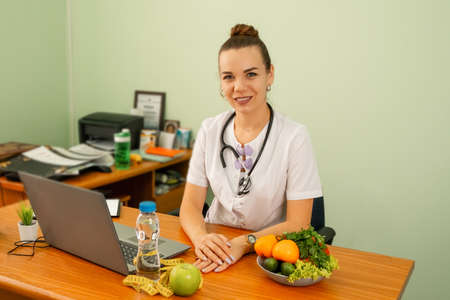 Happy smiling female dietitian in uniform with stethoscope at workplace. Young woman doctor sitting at the desk with eco food. Woman dietitian with tape measure working on a diet plan. Weight lossの写真素材