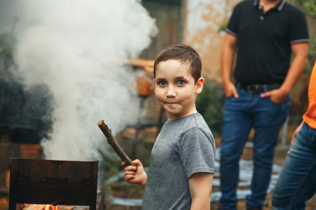 Child watching the fire. Barbecue in nature, group of preparing sausages on fire. Child boy playing with smoking wooden stick outdoors. Group of young friends camping and burning a wood fire.の写真素材