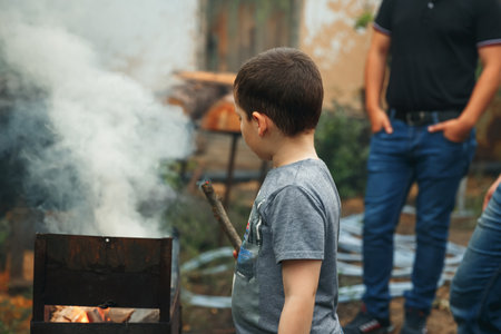 Child boy playing with smoking wooden stick outdoors. Child watching the fire. Barbecue in nature, group of preparing sausages on fire. Group of young friends camping and burning a wood fire.の写真素材