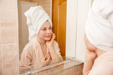 Young beautiful woman in bathroom. Portrait of young girl with towel on head in bathroom looks and touches her face in the mirror and enjoys youth. Natural beauty, home care for problem skin.の写真素材