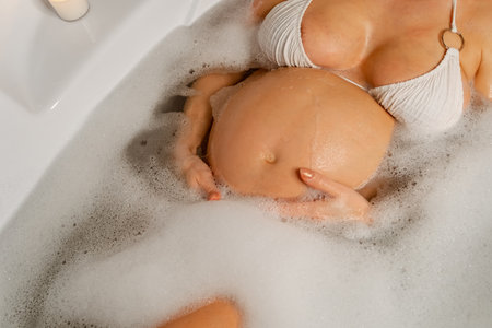 Close-up of a pregnant woman in a bubble bath, focusing on her belly. She is wearing a white bikini and enjoying a relaxing bath.の写真素材
