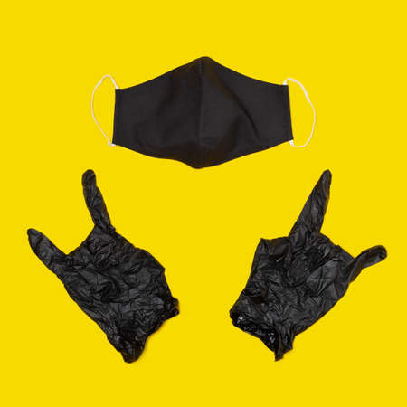 Black medical mask and black disposable surgical gloves making rock gesture on yellow background. Concept of hygiene and health protection during the epidemic covid-19.の写真素材
