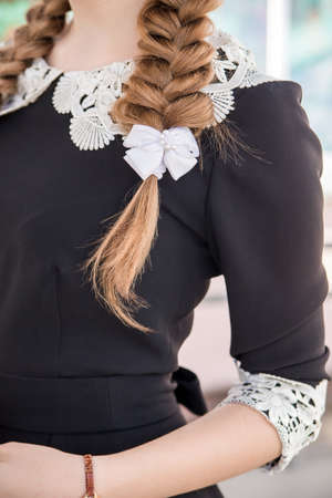 Braided hair with a white bow. A blonde schoolgirl in a strict dark school uniform with a white lace collar and cuffs. Elements of strict school uniform at the graduation and the last call.の写真素材