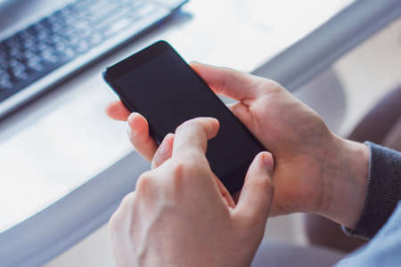 a photo of the hands of a young man wearing a gray hoodie and holding a black phone. Against a white...