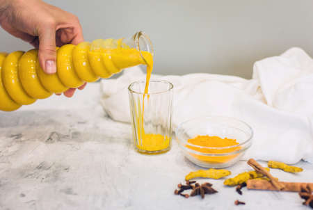 A woman's hand pours a turmeric latte with milk and cinnamon from a bottle into a glass. An elixir...
