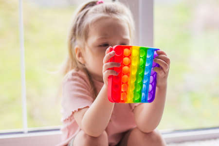 A little blonde girl sits near the window and plays with new trend sensory toy - rainbow pop . Antistress Ñolorful toy simple dimple. Squishy soft bubble toys rainbow colorの写真素材