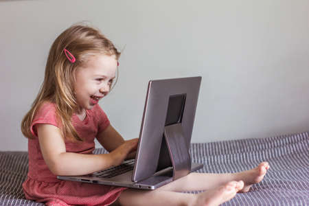 a small blonde girl in a red dress sits on the bed and looks at the laptop screen. Online communication, video communication, educationの写真素材