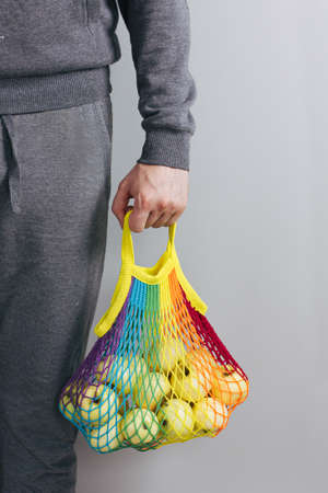 A man holds in his hands a reusable cotton shopping string bag, a multicolored rainbow with green apples inside. gray background. Zero waste conceptの写真素材