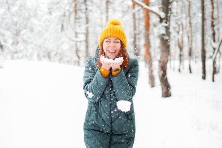 Cheerful girl with red hair in warm clothes playing with snow outdoors near the beautiful forestの写真素材