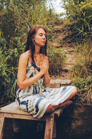 Woman practicing yoga on pier by river. Young Female wearing dress in lotus pose. Healthy Lifestyle...