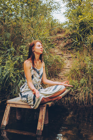 Woman practicing yoga on pier by river. Young Female wearing dress in lotus pose. Healthy Lifestyle and Yoga Concept.の写真素材