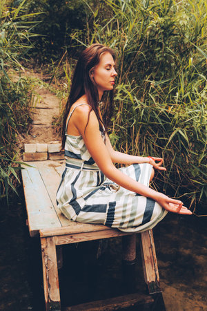 Woman practicing yoga on pier by river. Young Female wearing dress in lotus pose. Healthy Lifestyle and Yoga Concept.の写真素材