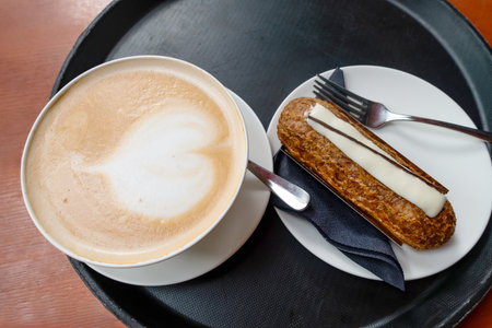 Latte art coffee cup alongside a delicious pastry on a plate, presented on a black tray in a warm...