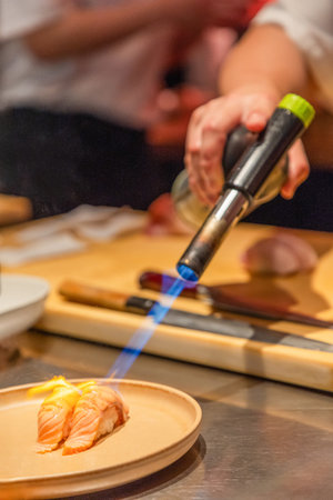 Culinary artist is using a torch to sear sushi on a wooden plate, showing skillful preparation in a vibrant kitchen environment.の写真素材