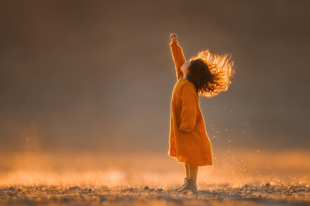 Young girl in an orange dress is raising her hand joyfully, surrounded by golden sunlight, capturing a moment of happiness and freedom.の素材
