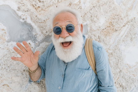 Elderly man with a white beard is smiling and waving cheerfully against a textured background, showcasing a joyful and friendly demeanor.の素材