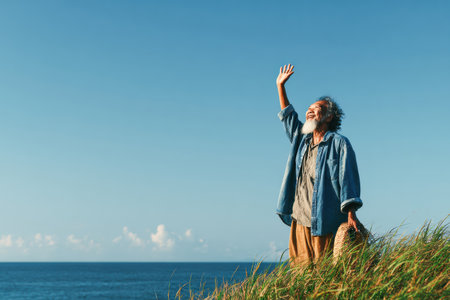 Elderly man is waving his hand while standing on a grassy hill, overlooking the ocean, with a bright sky and gentle breeze creating a serene atmosphere.の素材