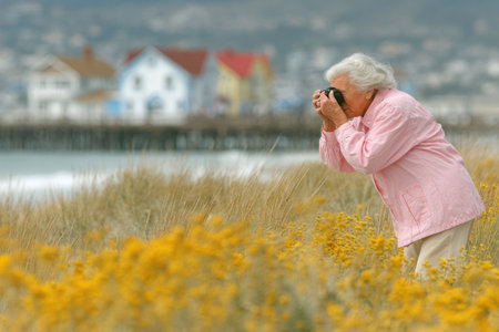 Elderly woman is capturing images of vibrant flowers by the beach, with colorful houses in the background, showing a serene outdoor moment.の素材