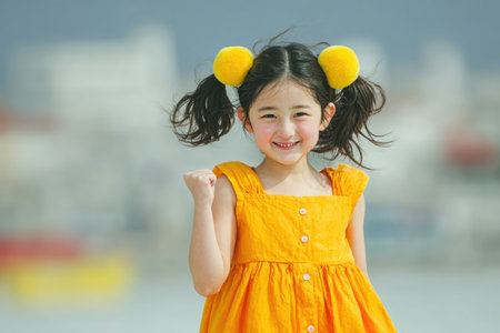 Young girl wearing a vibrant orange dress is smiling joyfully, showcasing playful hairstyle with pom-poms, capturing a cheerful moment outdoors.の素材