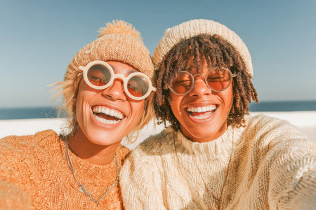 Two happy women are smiling brightly while wearing cozy sweaters and hats, enjoying a sunny day by the beach with clear blue skies.の素材
