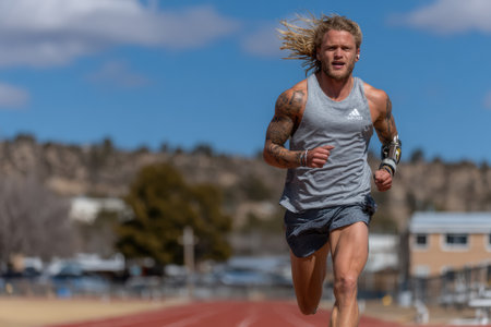 Athletic male runner is sprinting on a track, showing strength and determination, with a blurred background of trees and buildings.の素材