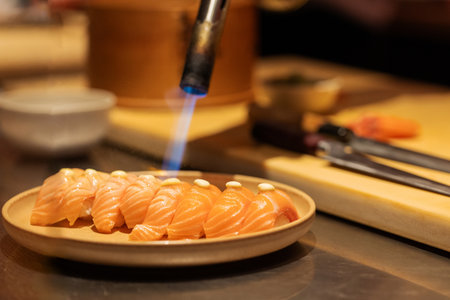 Sushi chef uses blowtorch on salmon fillets, showcasing culinary skills in a modern kitchen with utensils and ingredients visible.の写真素材
