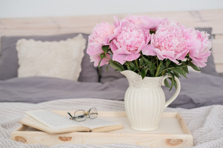 A vase with a bouquet of pink peonies, an open book and glasses on a tray on the bed.の写真素材