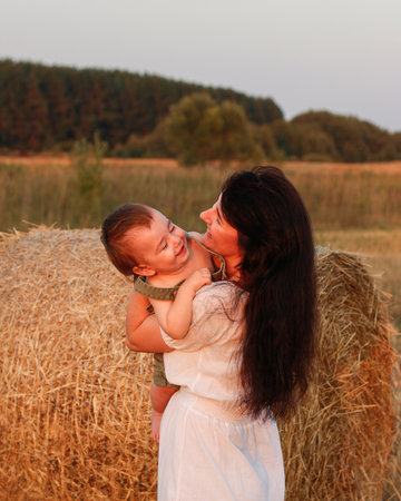 Mom with a baby in her arms in a field with bales of hay at sunset. Concept of happy family and summer vacationの写真素材