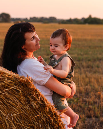 Mom with a baby in her arms in a field with bales of hay at sunset. Concept of happy family and summer vacationの写真素材