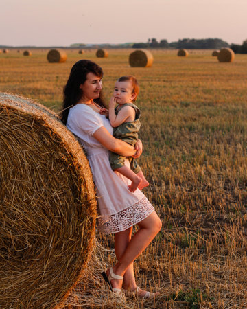 Mom with a baby in her arms in a field with bales of hay at sunset. Concept of happy family and summer vacationの写真素材