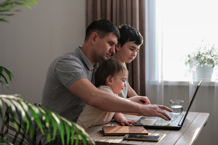 A man works at home with a laptop and looks after the kids. Father freelancer try to work at home with children. Home office and parenting at the same timeの写真素材
