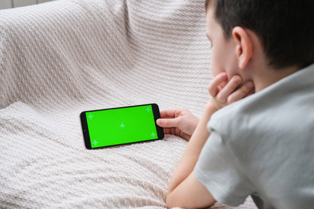 The boy looks at the screen of the phone with a chrome key indoors. An unrecognizable child uses a smartphone with a green screen to watch a video.の写真素材