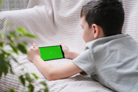The boy looks at the screen of the phone with a chrome key indoors. An unrecognizable child uses a smartphone with a green screen to watch a video.の写真素材