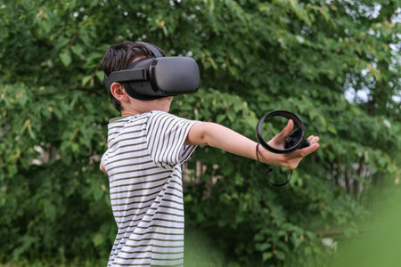 A seven-year-old boy in virtual reality glasses plays outside in the park. children using technologyの写真素材