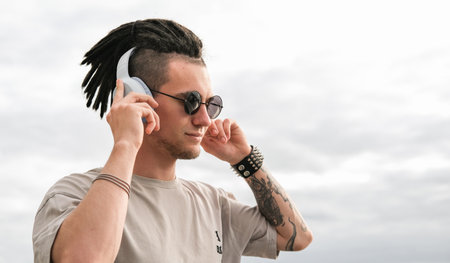 Portrait of a young guy with dreadlocks and piercings listening to music on headphones and enjoying momentの写真素材