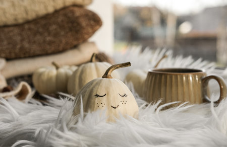 A white pumpkin with a cute painted face, a stack of knitwear and a cup of coffee on the windowsill. Hygge lifestyle, cozy autumn moodの写真素材