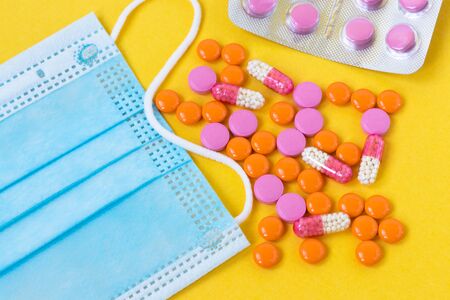 Multi-colored pills and a blue disposable mask on a bright yellow background, top view. Medication...