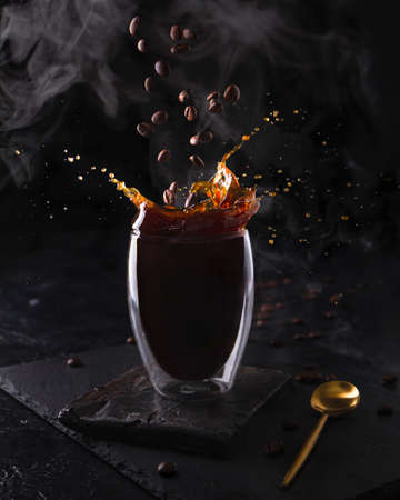 Hot espresso morning coffee in glass cup. Fragrant drink splashes with falling down coffee beans and steam on black background. Banner designの写真素材