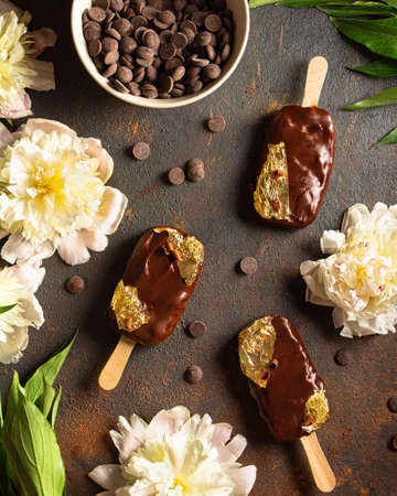 chocolate ice cream with edible gold on a stick on a dark background. Round pieces of chocolate around and in the bowl. Peonies and leaves. Top view. Chocolate ice cream made from dark, milk chocolateの写真素材