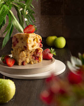 Two slices of charlotte pie on plates. Charlotte pie apple pie with strawberries. Powdered sugar and almonds are sprinkled on the charlotte pie. Tile wall in the background. Leaves and flowers of plants in a blur. Charlotte pie and apples on a wooden tableの写真素材