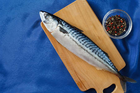 Raw mackerel fish whole, on a cutting board, bowl with a mixture of peppers and spices and a blue satin tablecloth. Top view with copy space.の写真素材
