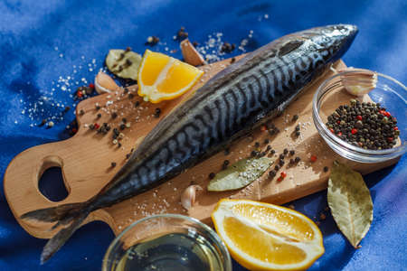 Mackerel fish raw on cutting board on blue satin tablecloth with ingredients for cooking, lemon,...