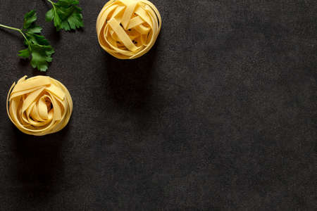 Fettuccine pasta, two raw uncooked nest and parsley - ingredients for an Italian dish, on dark background, top view, space to copy text.の写真素材