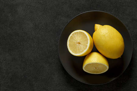Lemons whole and cut, on round plate on dark background, top view, space to copy text.の写真素材
