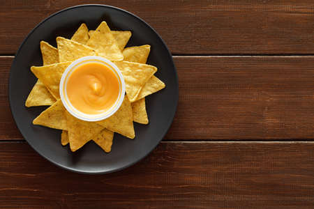 Nachos laid out on a plate with cheese sauce, on wooden plank dark background, top view, space to...
