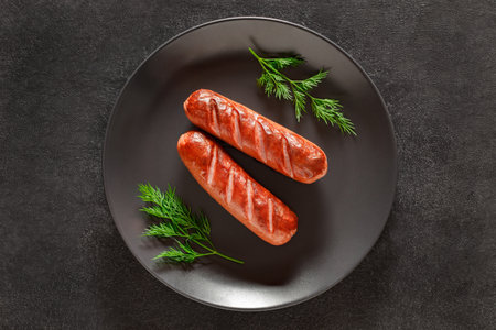 Two grilled sausages and springs of dill on dark background, top view, space to copy text